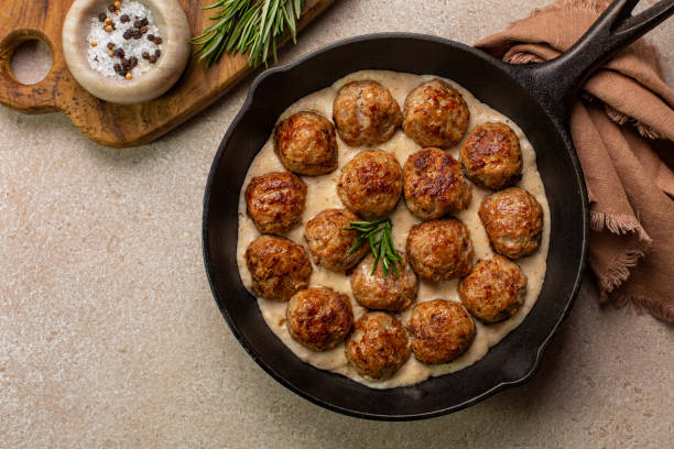 homemade swedish meatballs made with ground meat, onion, egg, bread crumbs and nutmeg. with creamy gravy in black pan skillet. on beige concrete table. - swedish meatballs stock pictures, royalty-free photos & images